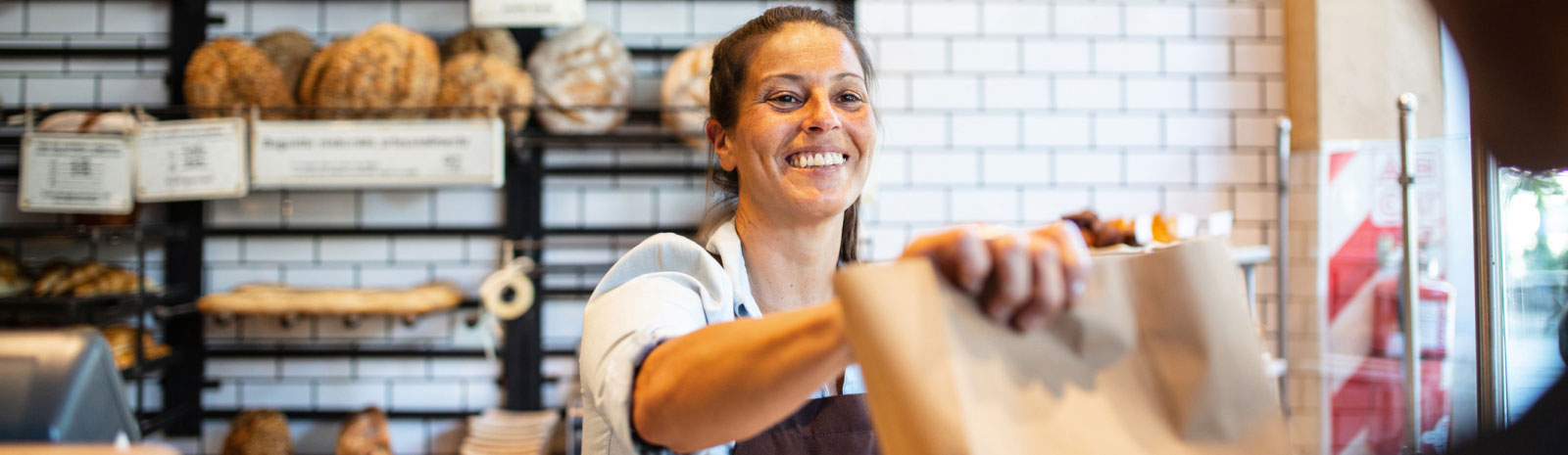 woman working at a bakery and handing a paper bag to someone out of frame